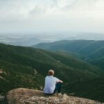 person sitting on the edge of a cliff over looking mountains during daytime